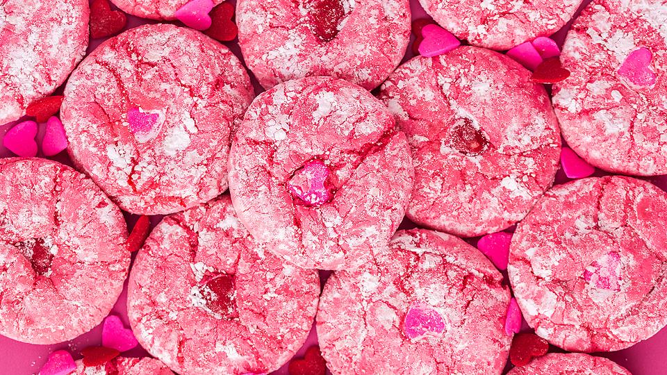 A close-up of several pink crinkle cookies topped with powdered sugar, surrounded by small pink and red heart-shaped candies.