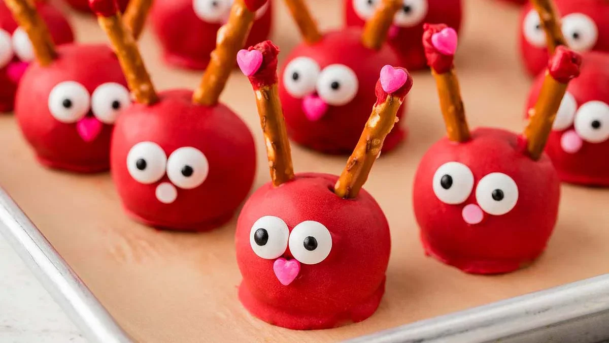 Red candy-coated treats shaped like reindeer with pretzel antlers, candy eyes, and pink heart noses, arranged on a baking tray.