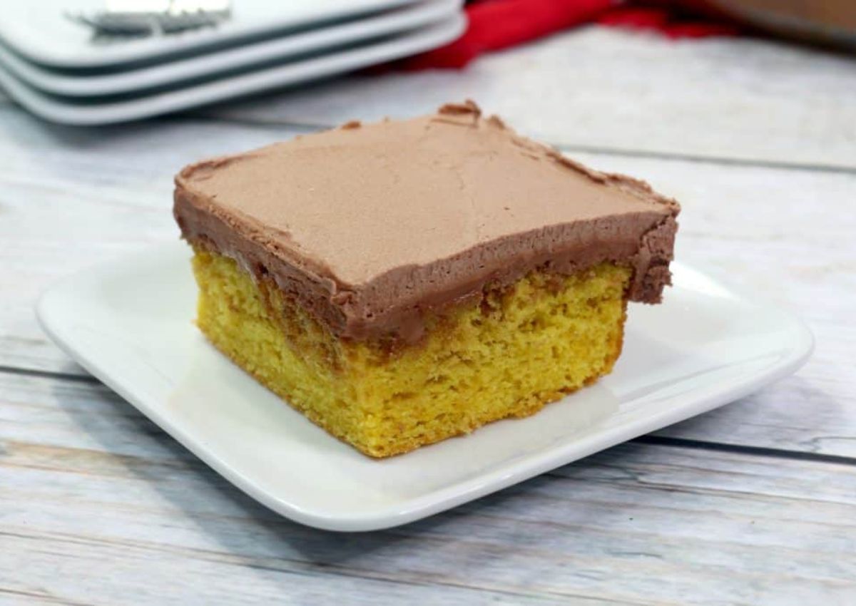 A square slice of yellow cake with chocolate frosting is served on a white plate.