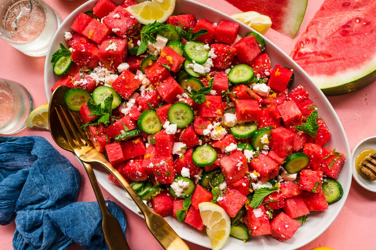 A platter of watermelon salad with cucumber slices, feta cheese, mint leaves, and lemon wedges, served with gold utensils on a pink background.