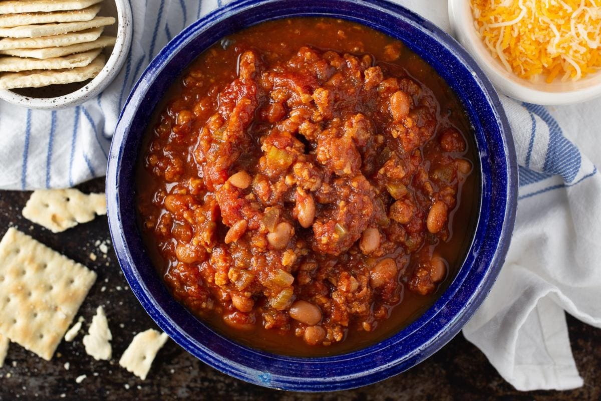 A blue bowl filled with chili containing ground meat, beans, and tomato sauce sits next to a bowl of shredded cheese and a plate of saltine crackers.