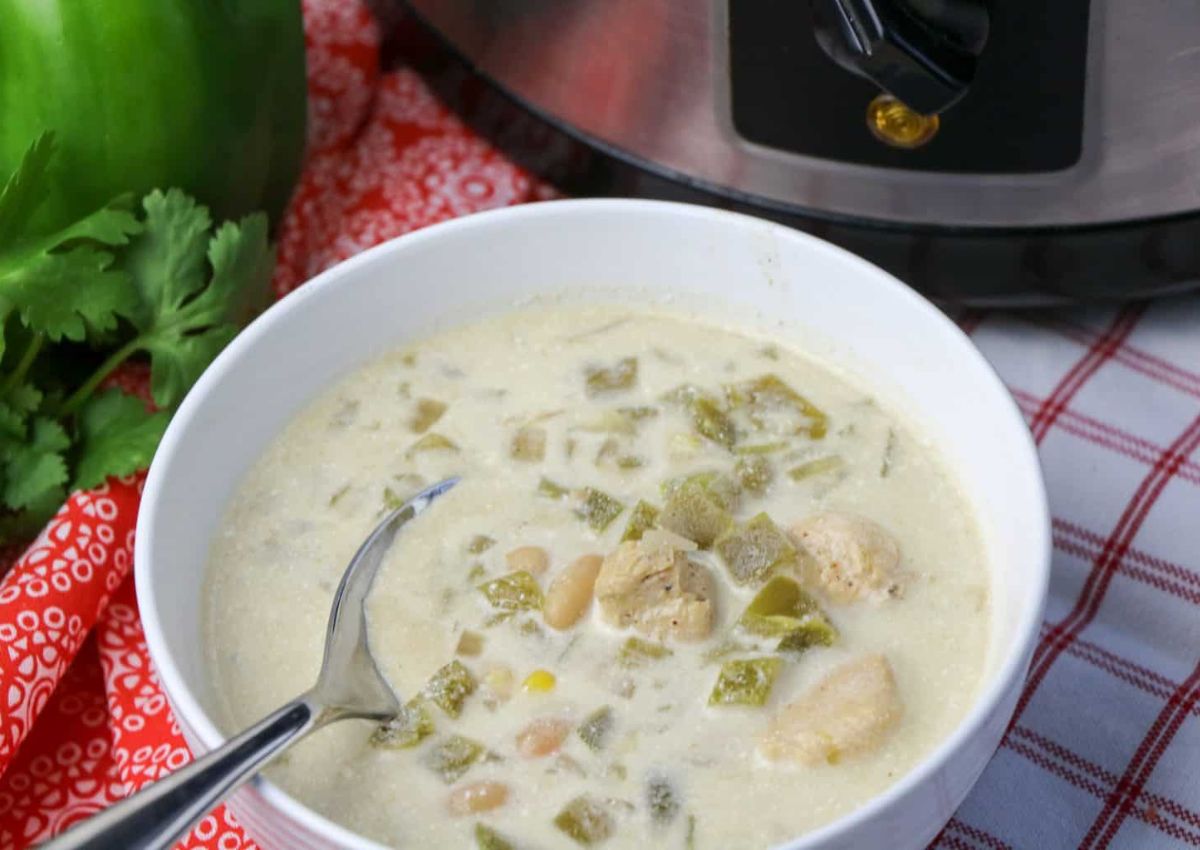 A bowl of creamy white chili with chunks of chicken, beans, and green peppers, with a spoon inside, placed near a slow cooker on a red and white checkered cloth.