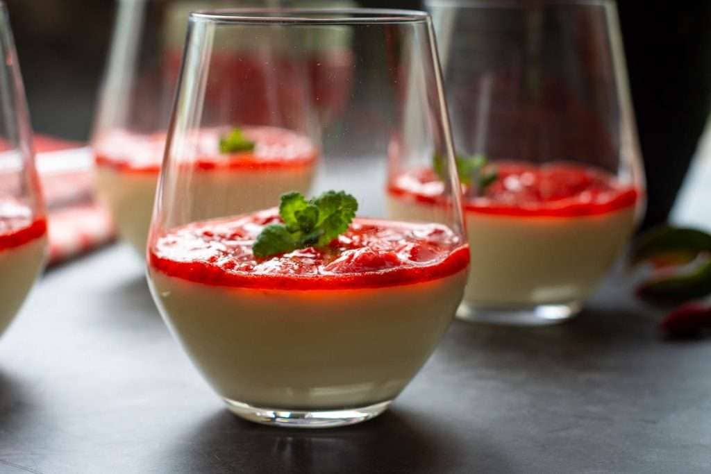 A glass of white panna cotta topped with red strawberry sauce and a small mint leaf sits on a dark surface, with similar desserts in the background.