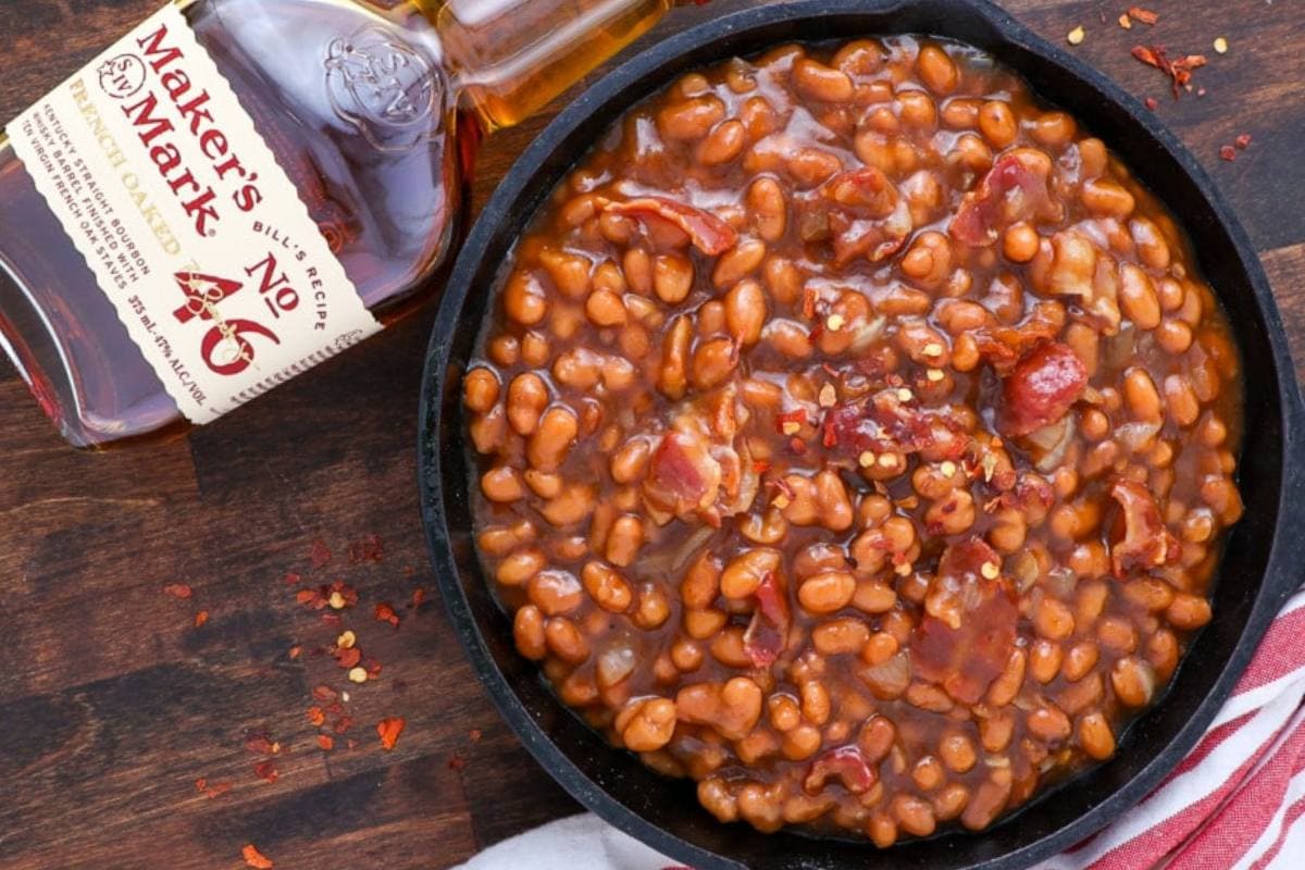 A skillet of baked beans with bacon sits beside a bottle of Makerโs Mark bourbon on a wooden surface.