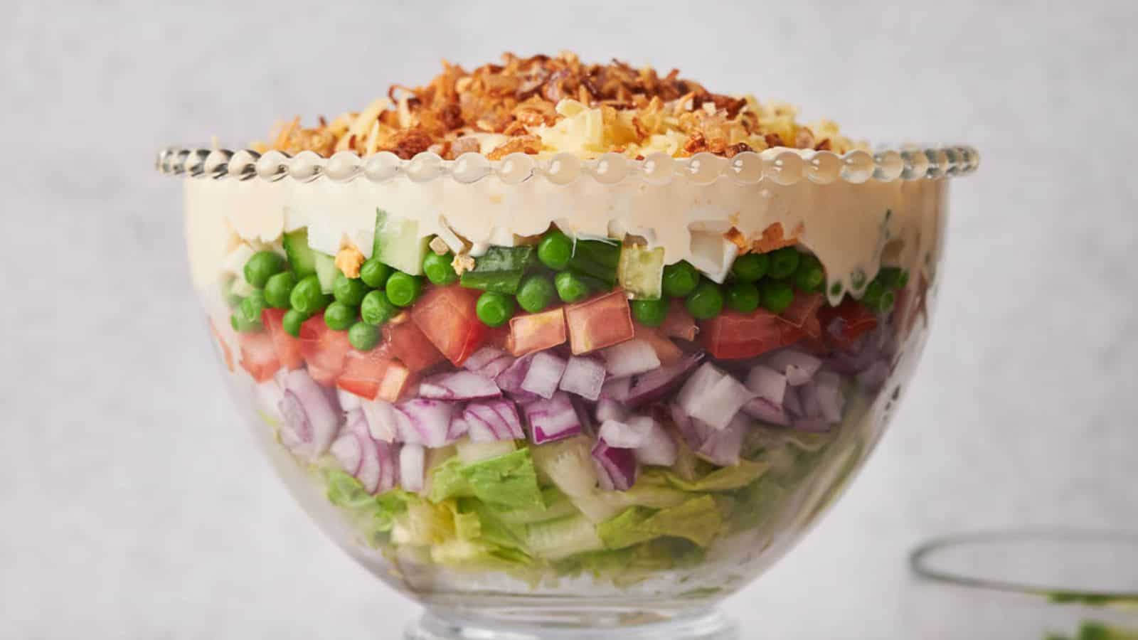 A glass bowl containing a layered salad with lettuce, red onion, tomato, peas, celery, mayonnaise, cheese, and a topping of crispy fried onions.