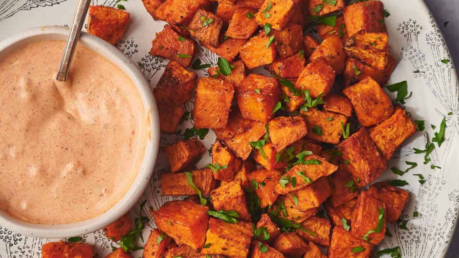A plate of roasted sweet potato cubes garnished with chopped herbs, served with a bowl of creamy dipping sauce.