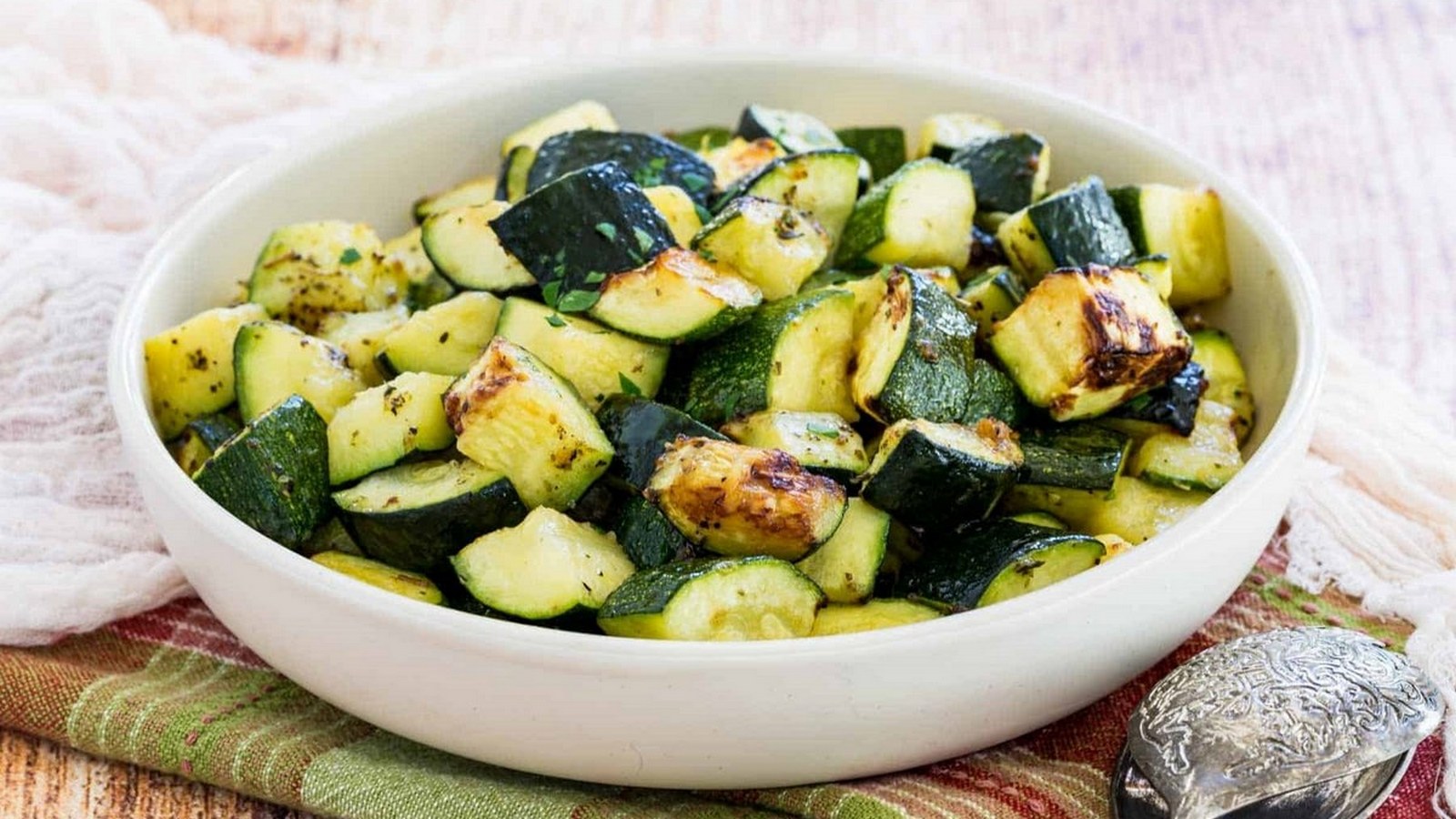 A white bowl filled with roasted zucchini pieces sits on a green and brown cloth next to a silver serving spoon.