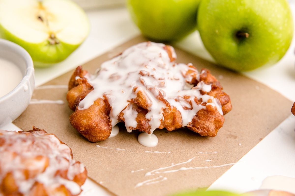 A glazed apple fritter sits on parchment paper, surrounded by green apples and a bowl of icing.