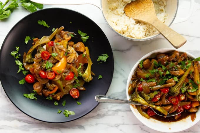 A plate of stir-fried vegetables with cherry tomatoes and herbs next to a bowl of white rice and a serving dish of the same stir-fry on a marble surface.