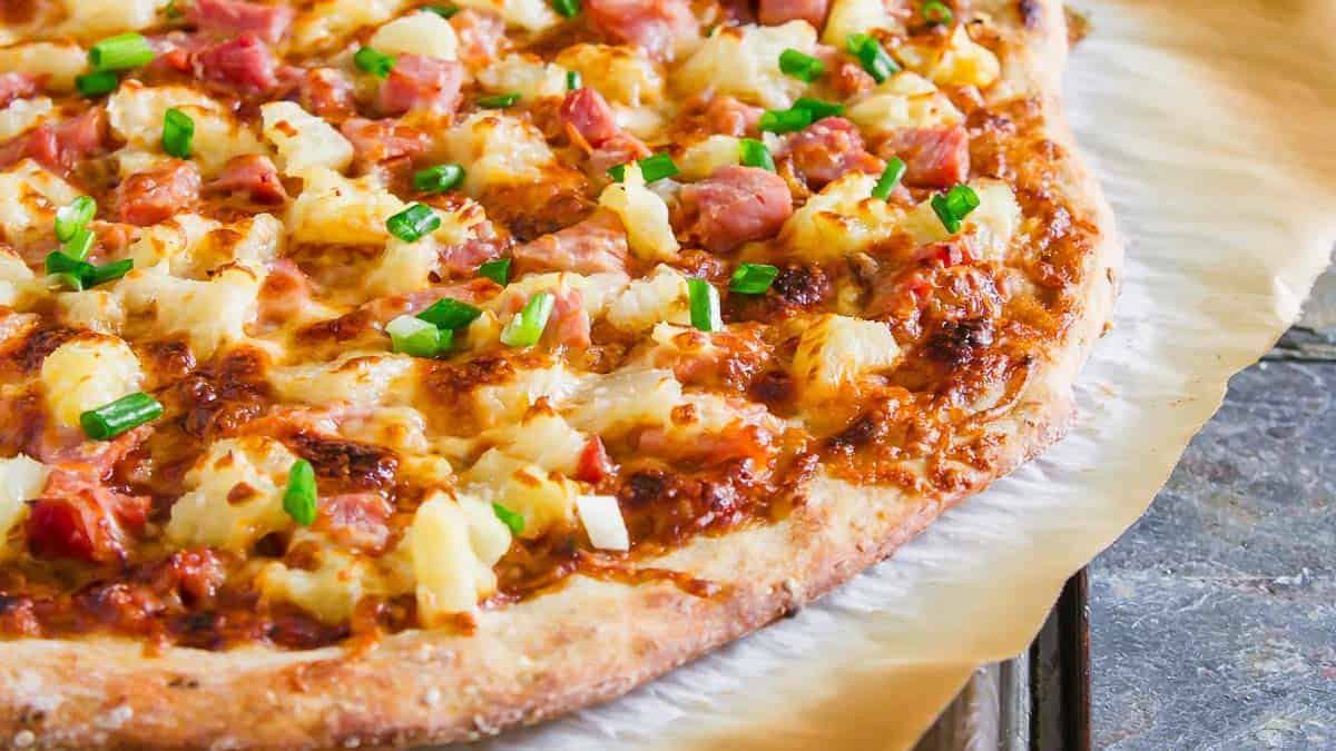 Close-up of a baked pizza topped with ham, pineapple chunks, melted cheese, and chopped green onions on a parchment-lined surface.