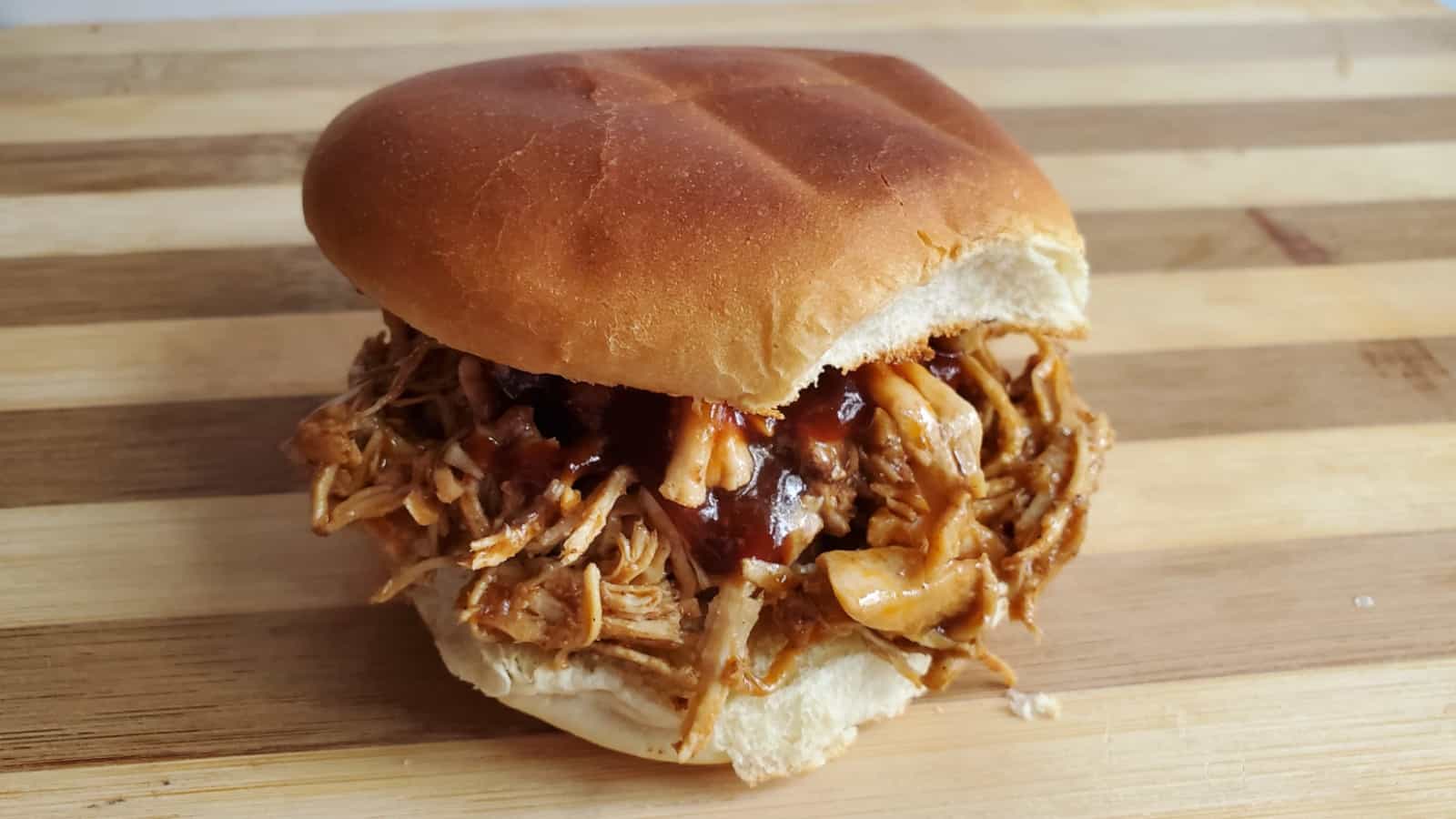 A pulled chicken sandwich with barbecue sauce on a soft bun, placed on a striped wooden cutting board.