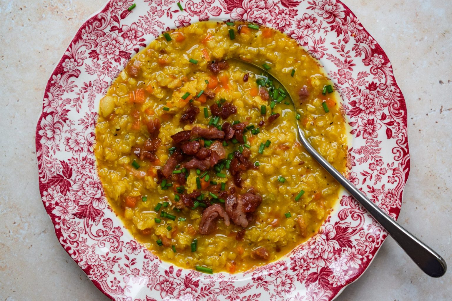 A bowl of yellow lentil soup with chopped vegetables and crispy bacon pieces, served on a red and white floral-patterned plate with a spoon.