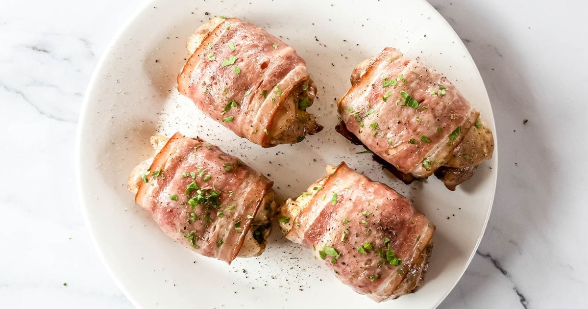 Four bacon-wrapped chicken breasts garnished with chopped herbs are arranged on a white plate, set against a marble background.