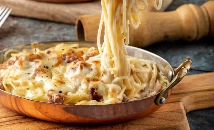 A pan of baked spaghetti with melted cheese and pieces of meat, with a fork lifting a portion, placed on a wooden board.