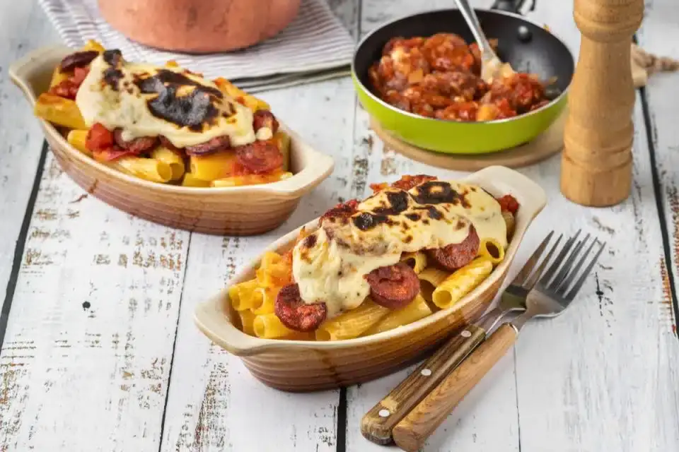 Two ceramic dishes filled with baked pasta topped with browned cheese and sausage slices, with a pan of tomato sauce and a pepper grinder in the background.