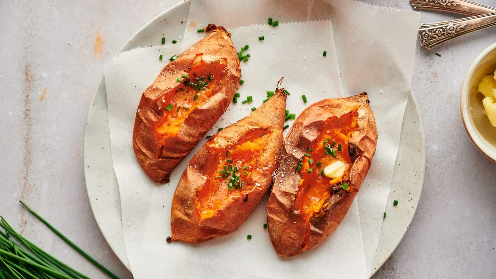Three baked sweet potatoes topped with chopped chives and a pat of butter are served on a round plate lined with parchment paper.