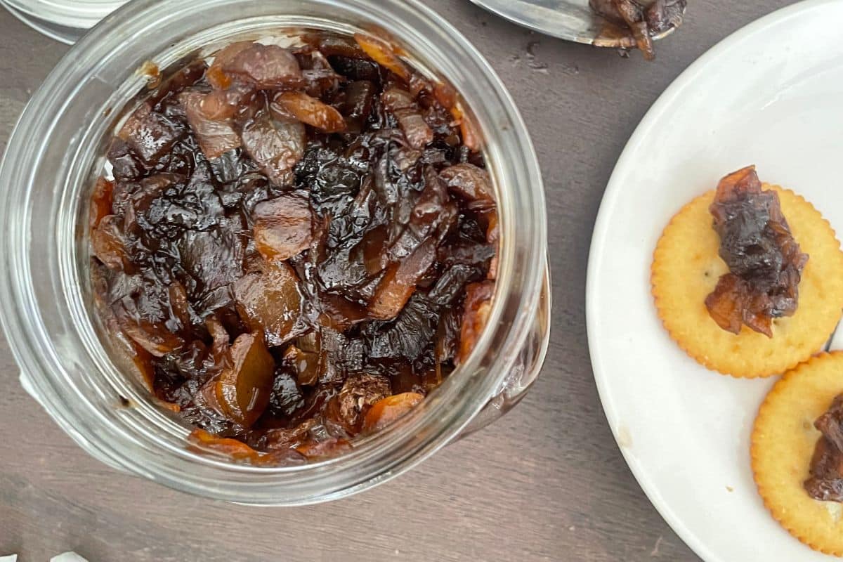 A jar of caramelized onion jam on a table next to a white plate with round crackers topped with the jam.