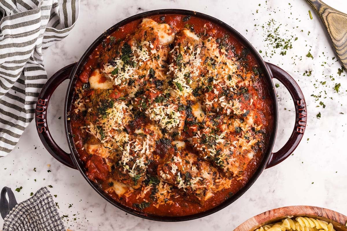 A round baking dish filled with baked chicken parmesan topped with melted cheese and herbs, placed on a marble countertop next to a striped towel and a wooden spoon.