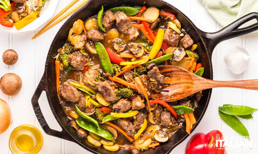 A cast iron skillet filled with beef stir-fry, mixed vegetables, and sauce, with a wooden spoon and fresh ingredients on a white table.