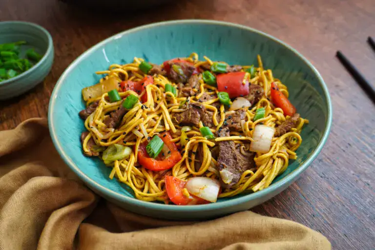A bowl of stir-fried noodles with beef, red bell peppers, onions, and green onions, served on a wooden table with chopsticks and a beige napkin.