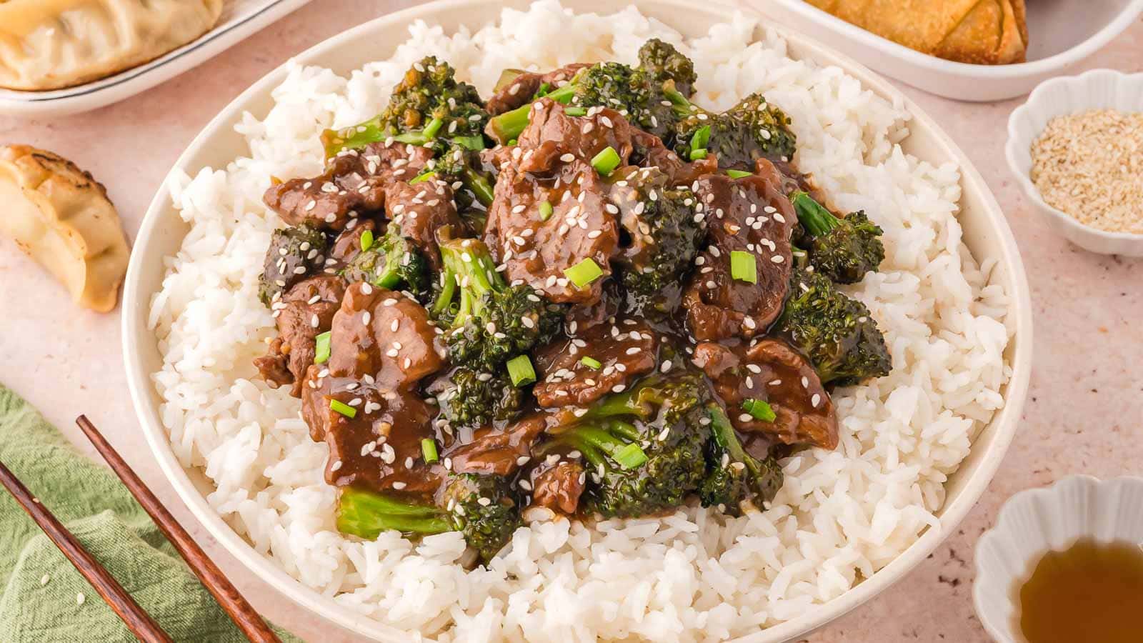 A bowl of white rice topped with beef and broccoli in sauce, garnished with sesame seeds and chopped green onions.