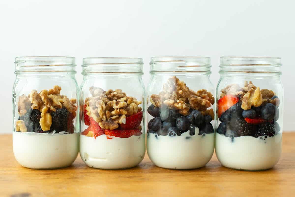 Four glass jars filled with yogurt, berries, and walnuts are arranged in a row on a wooden surface.