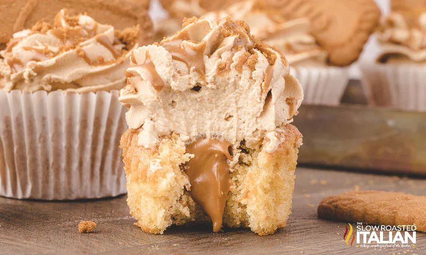 A cupcake with a creamy filling and swirled frosting is cut in half, showing the inside. Crumbled cookie pieces are sprinkled on top.