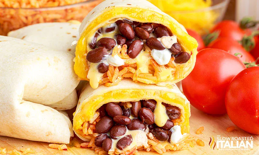 A burrito cut in half shows black beans, rice, cheese, and sour cream filling. Whole tomatoes and tortillas are in the background.