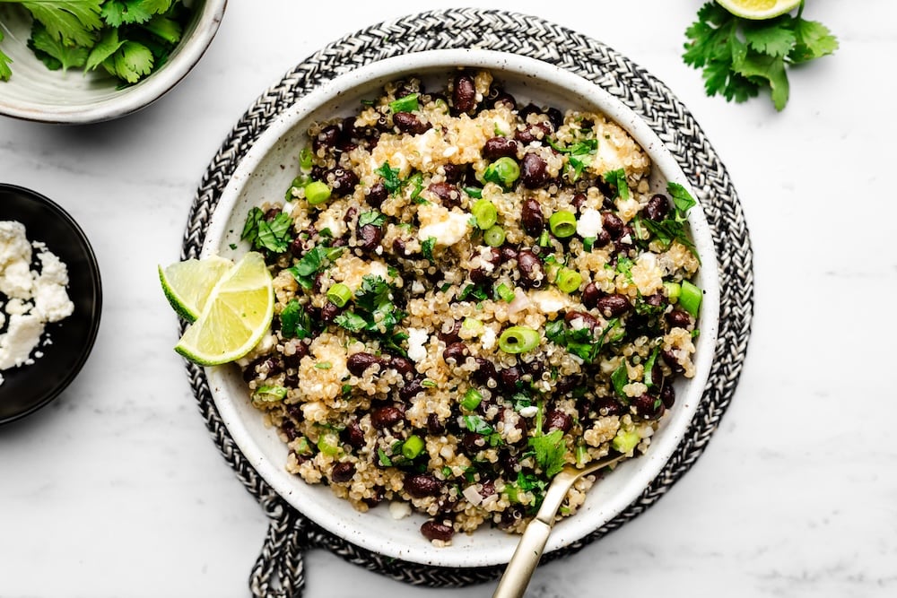 A bowl of quinoa salad with black beans, chopped green onions, cilantro, and lime wedges, served with a fork on a white surface.