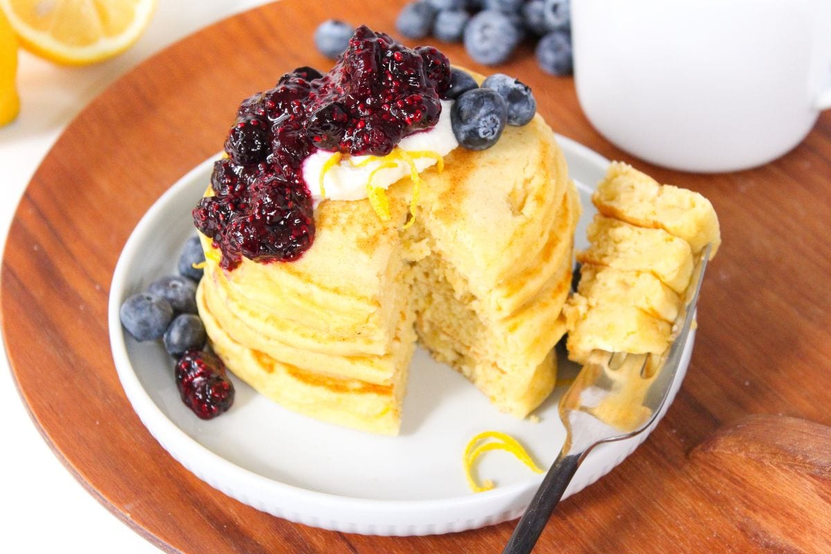 A stack of pancakes topped with berry compote, whipped cream, lemon zest, and blueberries sits on a white plate with a fork and extra blueberries on the side.