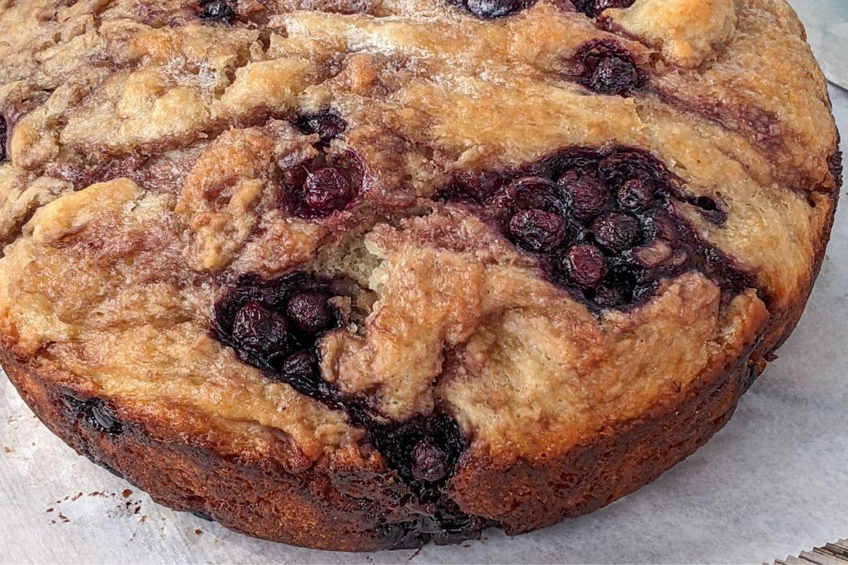 A close-up of a baked blueberry coffee cake with a golden-brown crust and visible clusters of blueberries on top.