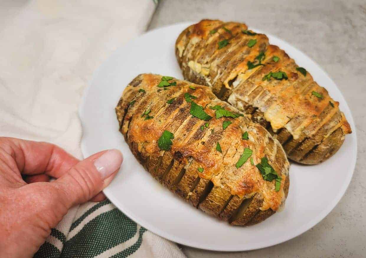 A hand holds a white plate with two baked hasselback potatoes topped with melted cheese and chopped parsley.