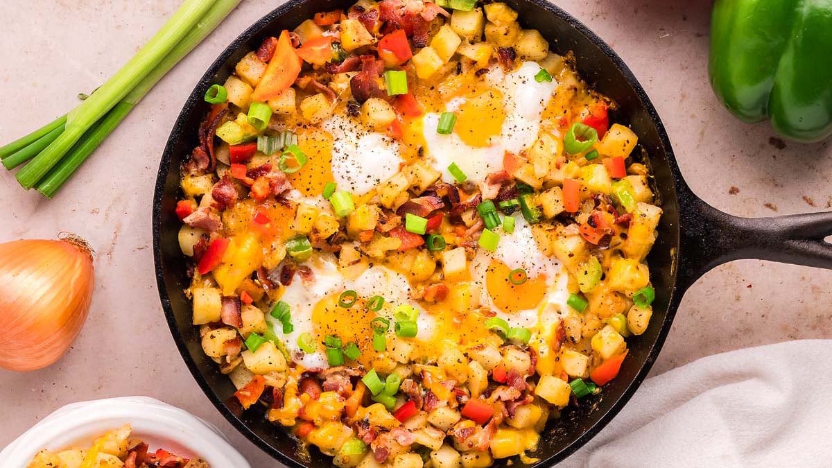 A skillet filled with a hash made of diced potatoes, eggs, cheese, peppers, onions, bacon, and chopped green onions.