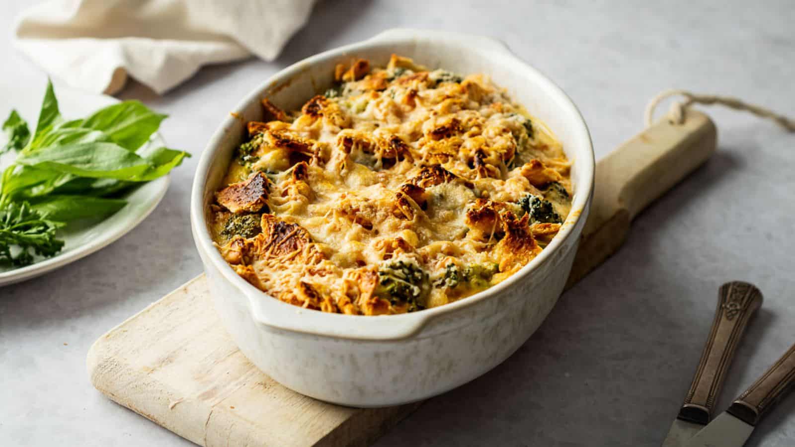 A baked casserole with melted cheese and bread pieces sits in a white oval dish on a wooden board, with fresh herbs on a plate nearby.