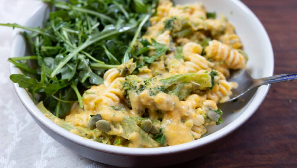 A bowl of rotini pasta with creamy yellow sauce, broccoli, and pumpkin seeds, served with a side of fresh arugula.