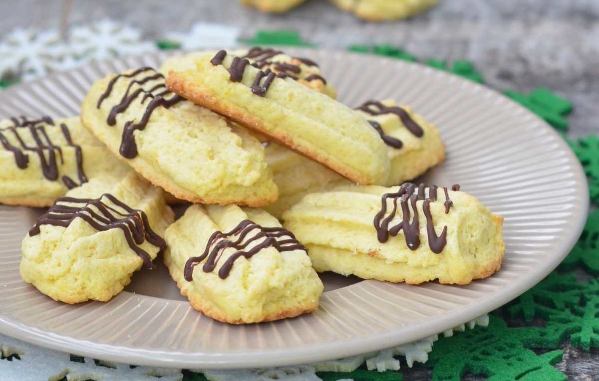 A plate of light yellow cookies, some drizzled with chocolate, arranged on a beige plate with green and white decorative placemats underneath.