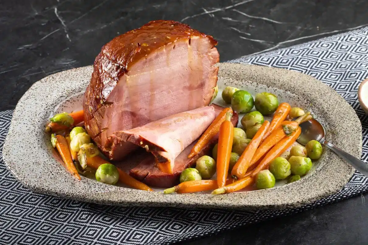 Glazed ham served on a stoneware platter with sliced carrots and Brussels sprouts, placed on a black and white patterned cloth.
