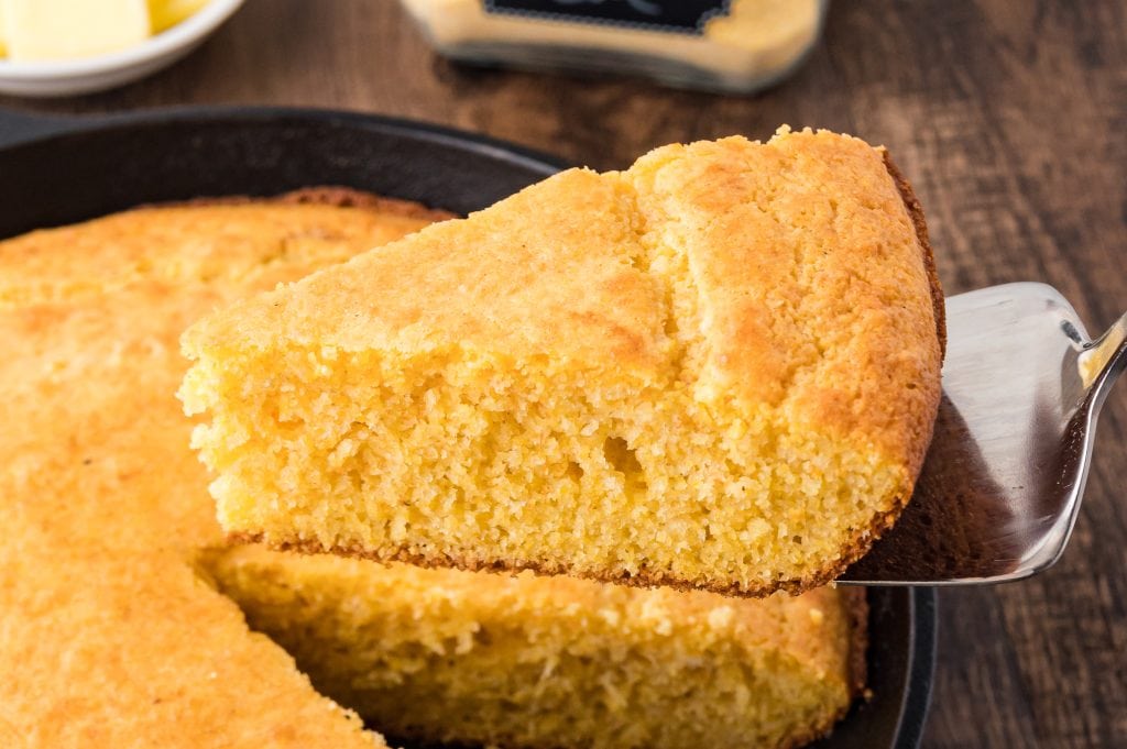 A close-up of a slice of cornbread being lifted from a cast iron skillet with a metal spatula.
