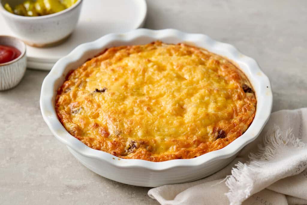 A baked cheesy casserole sits in a white fluted dish on a light surface, with a folded napkin beside it and small bowls in the background.