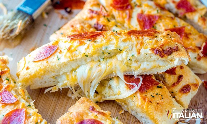 Cheesy breadsticks topped with melted cheese, slices of pepperoni, and herbs on a wooden surface, with cheese pulling between two breadsticks.