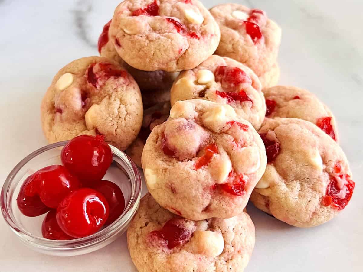 A stack of cookies with white chocolate chips and pieces of cherry, next to a small glass bowl filled with maraschino cherries on a white surface.