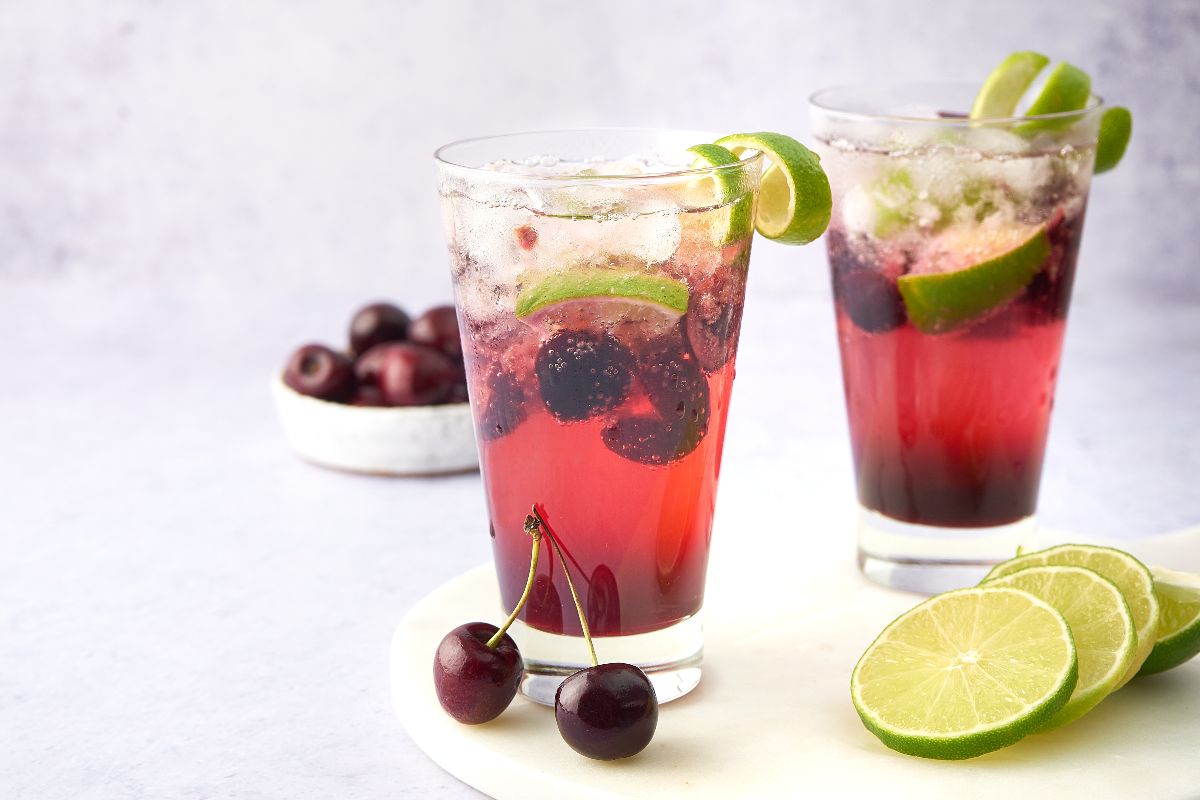 Two glasses of cherry and lime cocktail with ice, garnished with lime slices, are on a white surface with fresh cherries and lime slices nearby.