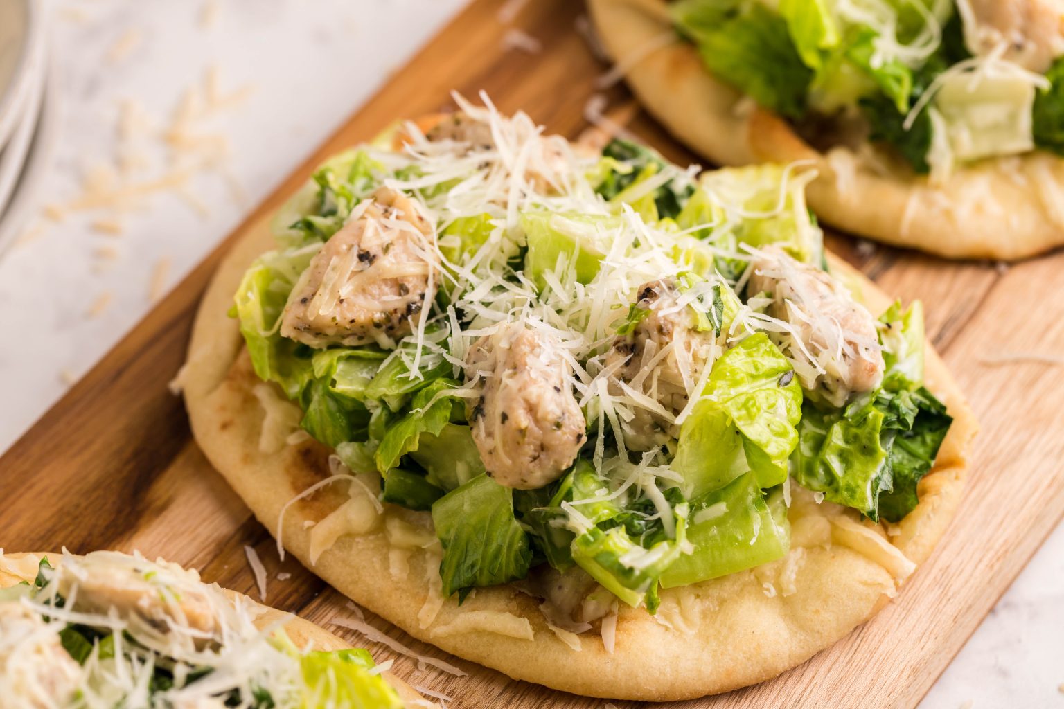 A flatbread topped with chopped romaine lettuce, grilled chicken pieces, and shredded parmesan cheese on a wooden board.