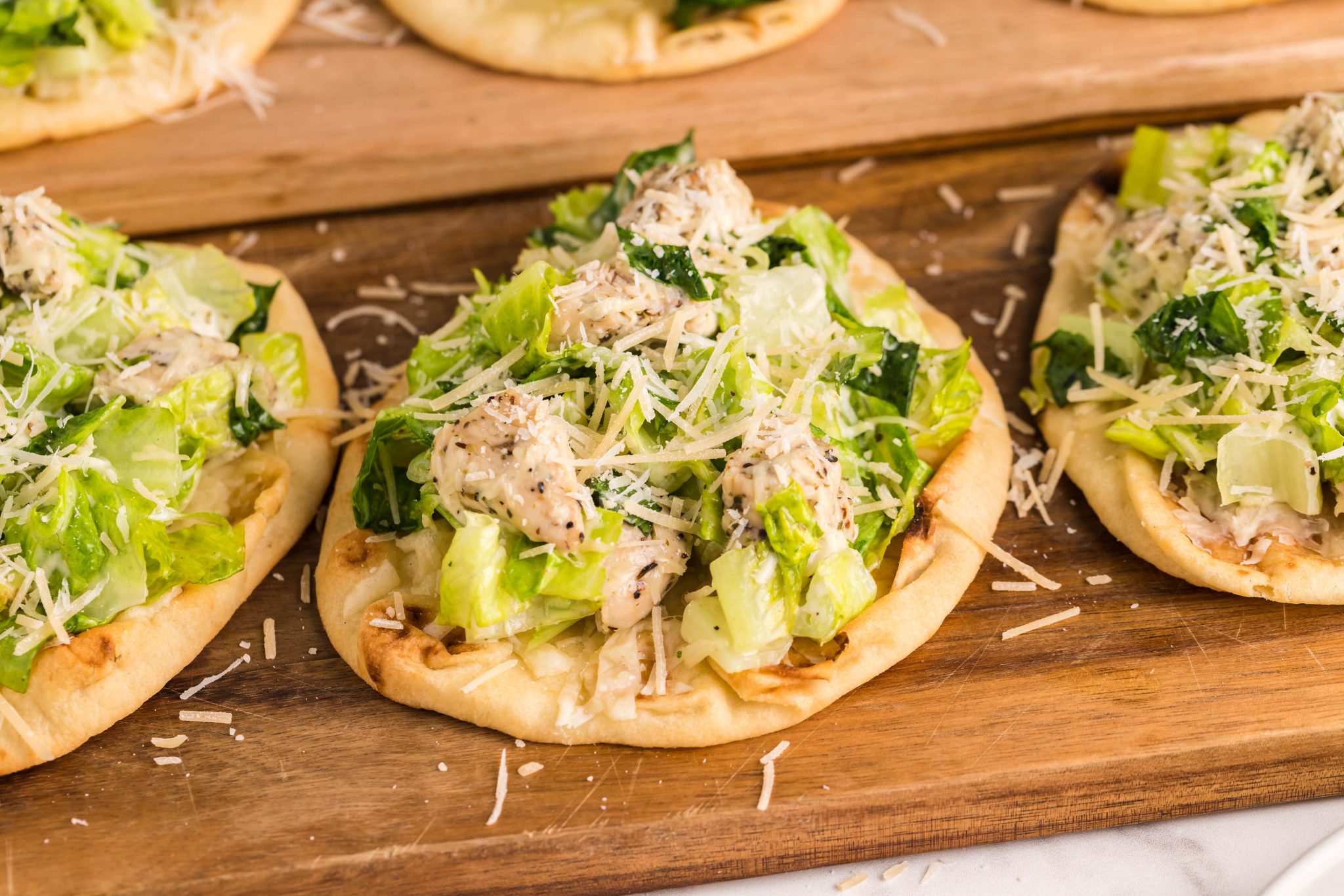 Flatbreads topped with chopped lettuce, seasoned chicken, and shredded cheese are arranged on a wooden serving board.