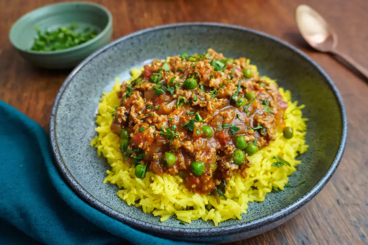 A bowl of yellow rice topped with a tomato-based sauce containing ground meat, peas, and herbs. A small bowl of chopped green herbs and a spoon are in the background.