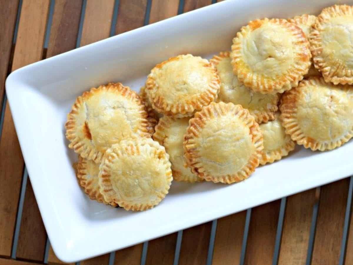 A white rectangular dish holds a pile of golden-brown, round hand pies with crimped edges, placed on a wooden surface.