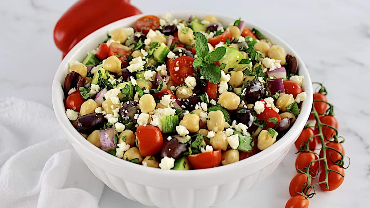 A white bowl filled with chickpeas, cherry tomatoes, olives, red onion, cucumbers, feta cheese, and fresh herbs sits on a white surface.