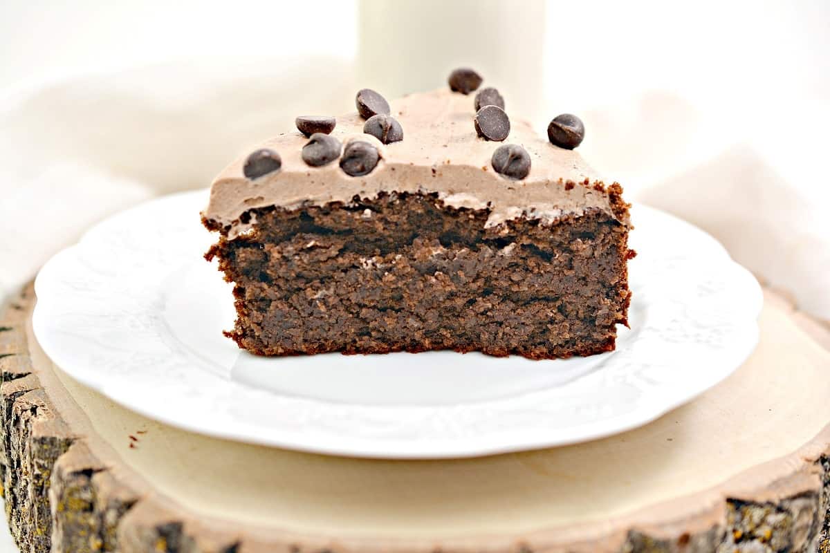 A slice of chocolate cake with frosting and chocolate chips on top, served on a white plate.