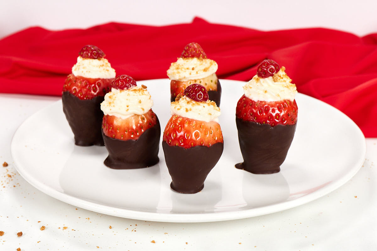 Five strawberries dipped in chocolate, topped with whipped cream and small berries, are arranged on a white plate with a red cloth in the background.