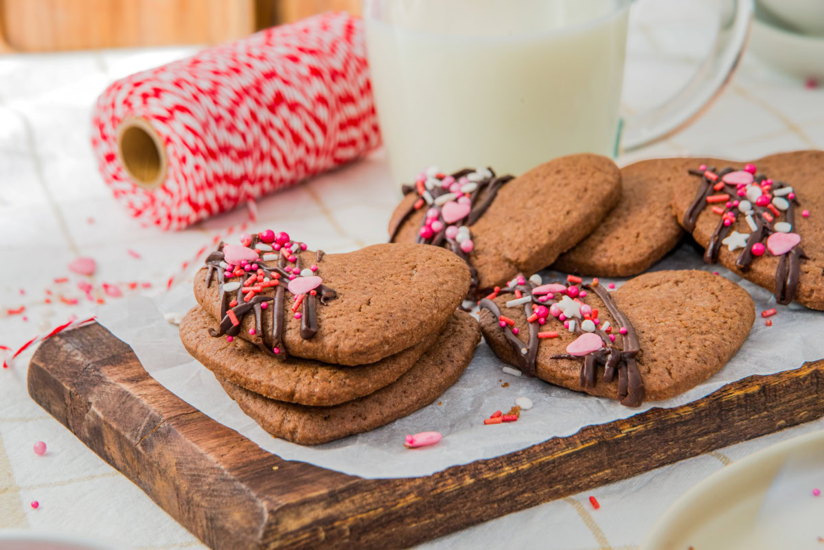 Heart-shaped cookies with chocolate drizzle and sprinkles are stacked on a wooden tray, with a spool of red and white twine and a glass of milk in the background.